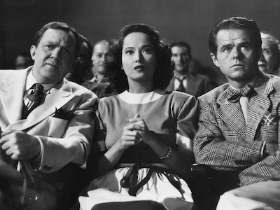 Merle Oberon sitting among a crowd.