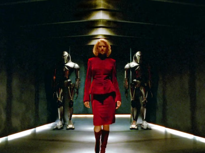 A woman in a red dress walking in front of two figures in armor.