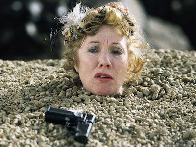 A woman buried up to her neck in the ground, with a gun near her head.