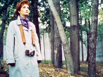 A woman standing among trees with a camera hanging from her neck.