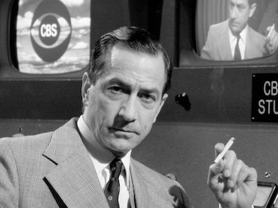 A news anchor smoking, with two TV monitors behind him. 