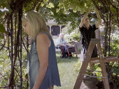 A person standing, a person trimming a vine, and a person sitting in a garden.