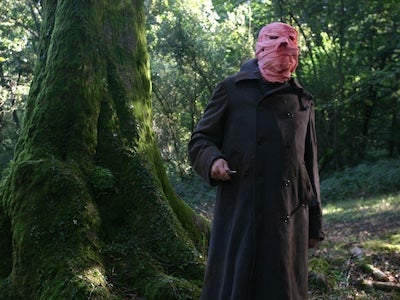 A person holding a knife in a forest, face wrapped in cloth.