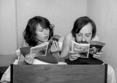 A woman and man looking at magazines in bed.