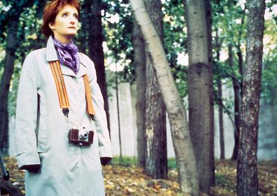 A woman standing among trees with a camera hanging from her neck.