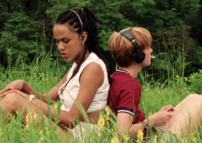 Two young women sitting in a field, sitting back to back.