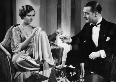 A man handing a cigarette to a woman, both dressed formally. 