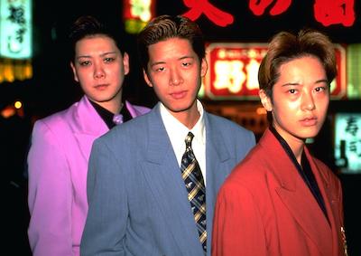 Three people in suits, neon signs in the background.