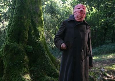 A person holding a knife in a forest, face wrapped in cloth.